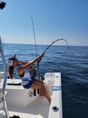 A woman on a boat is fishing, holding a bent rod over the water under a clear sky.