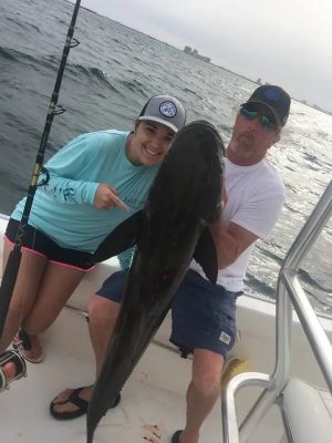 Two people on a boat display a large fish they caught, with ocean and distant buildings visible in the background.