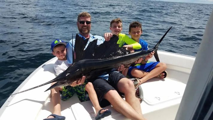 A man and three boys sit on a boat holding a large sailfish with the ocean in the background.