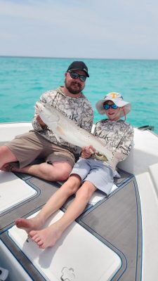 An adult and child sitting on a boat hold a large fish together, both wearing matching camo shirts and sunglasses.