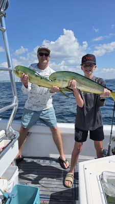 Two people on a boat hold a large mahi-mahi fish they caught, with blue ocean and sky in the background.