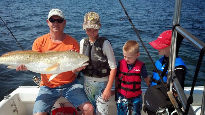 Four people on a boat; one adult holds a large fish while three children in life jackets look at it.