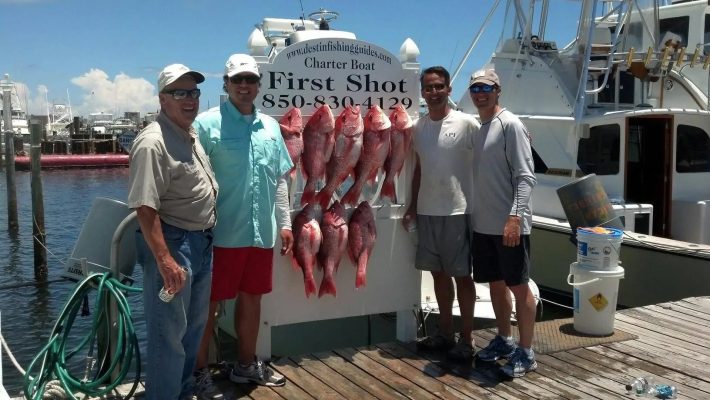 Four men stand on a dock, smiling beside a display of large red fish hanging in front of a charter boat sign.