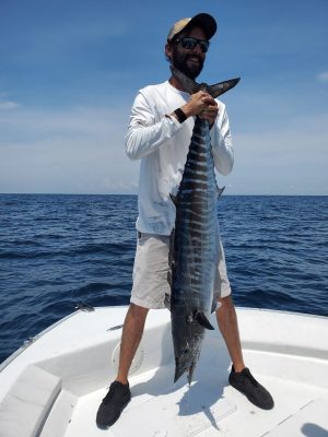 A man standing on a boat holds a large, striped fish vertically in front of him, with the ocean in the background.