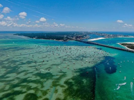 Aerial view of a coastal area with turquoise water, numerous boats, and a bridge connecting landmasses under a blue sky.